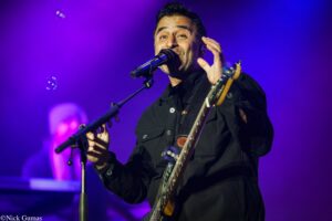 Male singer-guitarist performing on stage, singing into a microphone under purple lighting.
