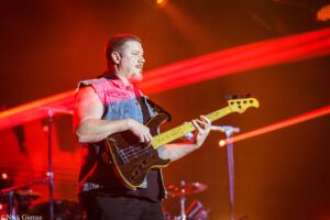 Male bass guitarist wearing a sleeveless denim jacket playing on stage under red lights during a live concert.