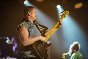 Male bassist in a sleeveless denim vest playing a dark bass guitar on stage with warm spotlights.