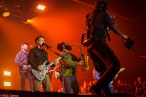 Band performing on a lit orange stage; guitarist front left sings into a microphone while a saxophonist plays nearby and a crowd watches.