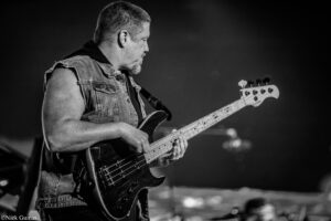 Side view of a guitarist in a sleeveless denim jacket playing a black electric bass on stage.