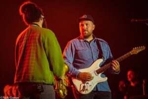 Two musicians on a red-lit stage: a guitarist in a blue shirt facing the camera with a white Fender Strat, and a saxophonist in a green plaid jacket seen from the back.