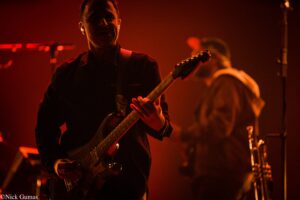 Guitarist in dark clothing playing an electric guitar on a dimly lit red stage with other band members blurred in the background.