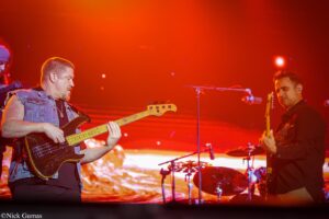 Two male guitarists performing on stage under orange-red lights, one playing a bass guitar and the other a regular electric guitar with a drummer in the background.