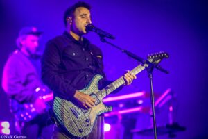 Male guitarist singing into a microphone while playing a striped electric guitar on a dimly lit stage with purple lighting.