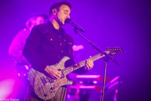 Male guitarist on stage, singing into a microphone while playing a striped electric guitar under purple lights.