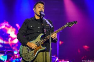 Male guitarist performing on a brightly lit stage, singing into a microphone while playing a striped electric guitar under blue and purple lights