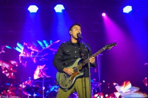 Male guitarist singing into a microphone on stage under blue and purple concert lighting