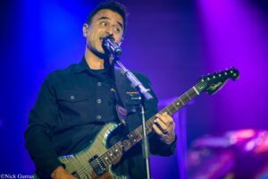 Male guitarist performing on stage, singing into a microphone under blue and purple stage lighting.