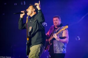 Male singer in a black shirt rocking a microphone on stage, blue lighting in the background with a guitarist in a denim vest behind him