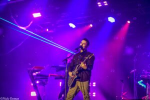 Male guitarist sings into a microphone on a purple-lit concert stage with bubbles and keyboard gear in the background, creating a vibrant live performance scene.