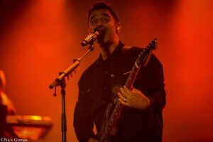 Male singer-guitarist on stage under orange lighting, singing into a mic while playing a bass guitar./