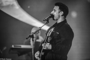 Male guitarist singing into a microphone on stage, wearing an in-ear monitor and dark jacket, black-and-white concert photo