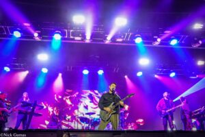 Band performing on a colorful stage with blue and purple lights; lead guitarist and singer center stage.