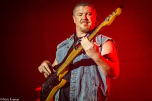 Male bass guitarist in a sleeveless denim vest plays a sunburst bass on a red-lit stage, looking focused.