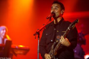 Male musician singing into a microphone while playing a bass guitar on stage under warm orange-red lights.
