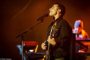 Male guitarist singing into a microphone on stage, wearing a dark shirt, with warm orange stage lighting.