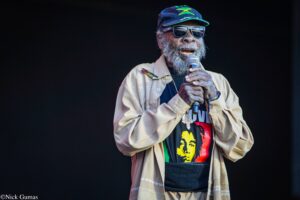 Reggae singer with gray beard and Jamaica flag cap, wearing sunglasses, holding microphone on stage.