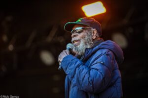 Man with gray beard wearing a blue jacket and sunglasses sings into a handheld microphone on stage at night, orange stage light behind him.