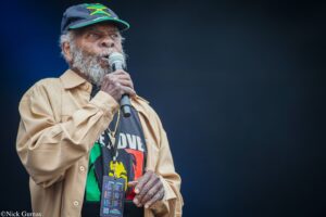 Older man with gray beard singing into a microphone on a dark stage, wearing a Jamaica flag cap and a beige jacket.