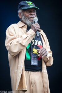 Elderly man with gray beard wearing a Jamaica flag cap speaks into a microphone on stage, beige jacket, and black shirt with a colorful design.
