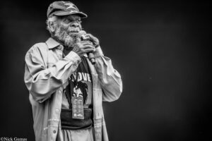 Older man with a gray beard wearing a cap and light jacket, singing into a handheld microphone on stage.