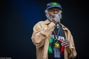 Male reggae singer with gray beard in a Jamaica flag cap and tan shirt, singing into a microphone on stage.
