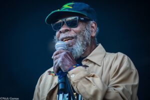 Bearded man in a navy cap and sunglasses singing into a microphone on stage.