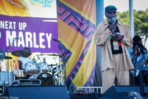 Male performer with microphone on a colorful outdoor stage; beige outfit, hat and sunglasses, badge around neck, drums and colorful backdrop behind him.