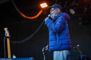 Male singer with gray beard in a blue puffer jacket and cap, singing into a handheld microphone on stage, guitar visible to the left.