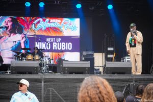 Stage performance with a man singing on the right, microphone in hand, and a large colorful screen in the background showing 'California Roots' and 'Niko Rubio' (2026).