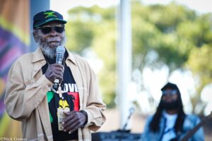 Older Black man with gray beard wearing a Jamaica flag cap and sunglasses, speaking into a microphone outdoors.