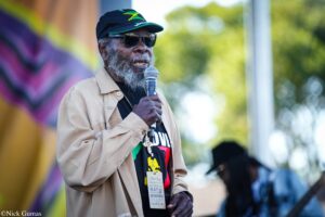 Elderly man with gray beard, Jamaica-flag cap, sunglasses, and beige jacket, speaking into a microphone on stage.