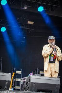 Older man with gray beard singing into a microphone on a stage, wearing a beige outfit and a Jamaican-themed shirt and cap amid blue stage lights.