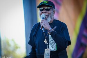 Older man wearing sunglasses and a Jamaican flag cap sings into a microphone on stage at a festival.