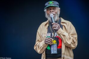 Performer with gray beard singing into a handheld microphone on stage, wearing a Jamaican flag cap and beige jacket.