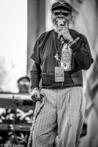 Black-and-white photo of an older man singing into a microphone on stage, wearing a cap, sunglasses, and a festival badge around his neck, holding a patterned cane/prop.