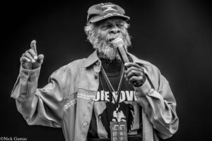 Older man with gray beard singing into a microphone on stage, wearing a cap and light jacket, gesturing with his left hand.