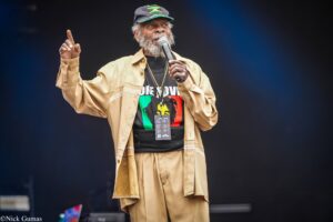 Male reggae singer on stage in a tan outfit, wearing a cap with the Jamaican flag, speaking/singing into a microphone while gesturing with his raised finger on a dark background.