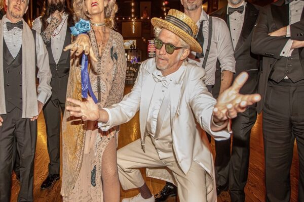 Group of guests in vintage attire at a party, man in a white suit and straw hat reaches toward the camera.