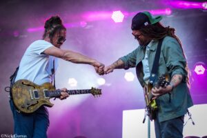 Two guitarists on a colorful stage give a fist bump mid-performance, each holding an electric guitar under purple stage lights.