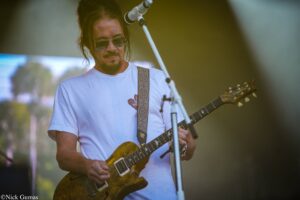 Guitarist wearing sunglasses and a white T-shirt plays a sunburst electric guitar on stage.