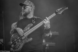 Bass guitarist on stage wearing a camo cap and sunglasses, focused on playing a patterned bass guitar.