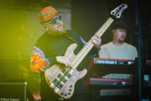 Bass guitarist wearing an orange camo hat and sunglasses plays a wooden bass on stage, with a keyboardist in the background under yellow lighting.
