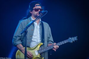Musician wearing a backwards cap and sunglasses plays a yellow electric guitar on a blue-lit stage at a live performance with a microphone nearby.