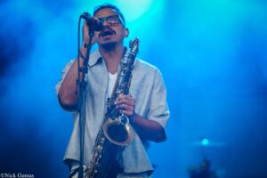 Male saxophonist wearing sunglasses sings into a microphone on stage under blue lights, holding his saxophone close.