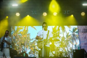 Male guitarist in a white t‑shirt performing on a smoky, yellow-lit stage with a tropical palm backdrop; a saxophonist is visible to the left and instruments surround the scene.