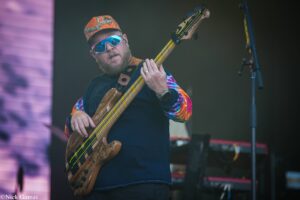 Bass guitarist performing on stage, wearing a camo hat and blue reflective sunglasses with tie-dye sleeves.