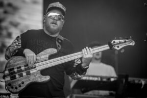 Bass guitarist wearing a cap and mirrored sunglasses, playing a striped bass on stage.