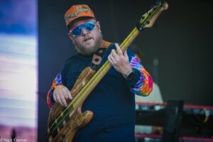 Male bass guitarist on stage wearing an orange camouflage cap and blue mirrored sunglasses, playing a wooden bass with neon-green strings.